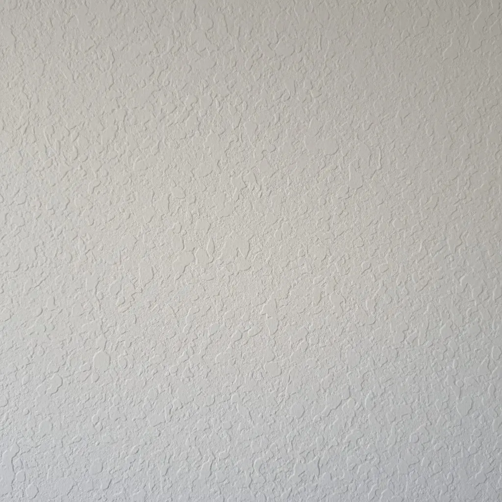 A close view of knockdown texture on a finished drywall wall, the flattened spatter pattern raking across the surface under a low light so the relief reads.