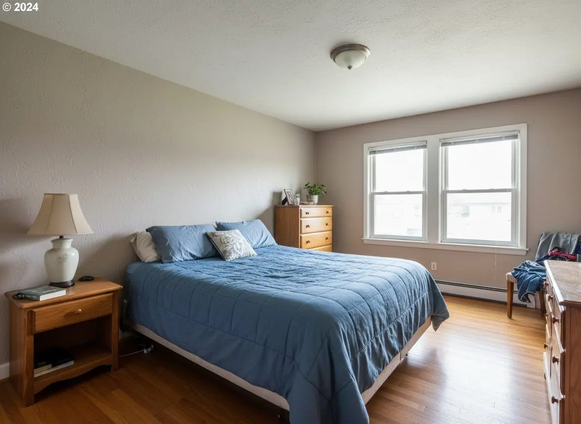 A bright, finished, furnished bedroom in a well-kept home, the walls and ceiling evenly textured and painted, with a made bed and a wooden floor